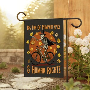 May include: A black garden flag features a skeleton riding a bicycle, holding a jack-o'-lantern. The flag has the text "BIG FAN OF PUMPKIN SPICE & HUMAN RIGHTS" in orange. The design includes orange leaves and flowers, set against an orange circle.