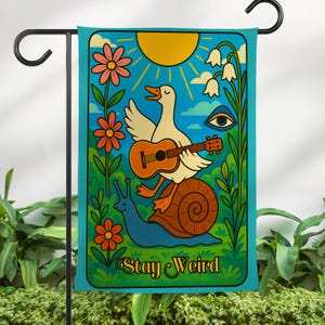 May include: A whimsical garden flag featuring a cartoon duck playing a ukulele while riding a snail. The design includes a sun, flowers, and the text "Stay Weird" in a retro style. The flag is turquoise with a black metal pole.