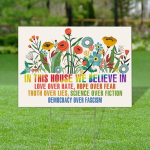 May include: A rectangular yard sign with a floral design and the text "IN THIS HOUSE WE BELIEVE IN" in rainbow colors. The sign also includes the phrases "LOVE OVER HATE, HOPE OVER FEAR, TRUTH OVER LIES, SCIENCE OVER FICTION, DEMOCRACY OVER FASCISM."