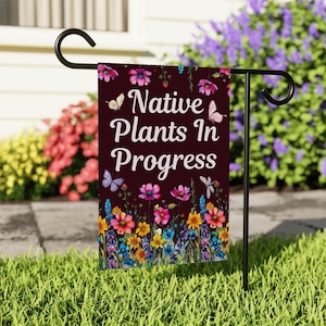 May include: A garden flag with the text "Native Plants In Progress" on a dark maroon background. The flag features colorful flowers and butterflies. The flag is displayed on a black metal stand in a grassy yard.