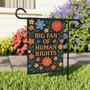 May include: A garden flag with the text "BIG FAN OF HUMAN RIGHTS" in gold lettering, surrounded by colorful flowers and green leaves on a navy blue background. The flag is displayed on a black metal stand in a garden setting.