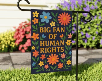 Puede incluir: Una bandera de jardín azul marino con el texto dorado "BIG FAN OF HUMAN RIGHTS", rodeada de flores coloridas y hojas verdes. La bandera se muestra en un soporte de metal negro en un jardín.