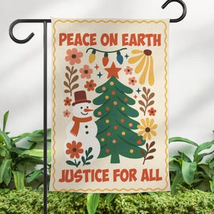 May include: A decorative garden flag with the words "PEACE ON EARTH" and "JUSTICE FOR ALL" in red. The flag features a snowman, Christmas tree, flowers, and string lights in a colorful, festive design. The flag is displayed on a black metal stand.
