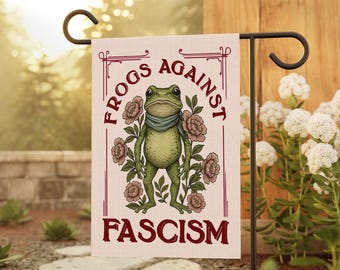 May include: A garden flag featuring a cartoon frog wearing a scarf, surrounded by flowers. The flag reads "FROGS AGAINST FASCISM" in maroon text within a decorative border. The flag is displayed on a black metal stand in a garden setting.