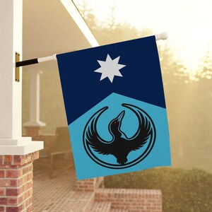 Op de afbeelding: Een rechthoekige vlag met een donkerblauw bovenstuk en een lichtblauw onderstuk. De vlag heeft een witte ster en een zwart vogel-embleem met een rood oog, geplaatst in een zwarte cirkel. De vlag is gemonteerd op een witte paal.