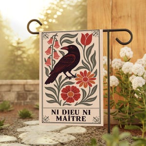 May include: A garden flag featuring a dark red raven perched among red flowers and green leaves. The flag has the text "NI DIEU NI MAÎTRE" in black. The flag is displayed on a black metal stand in a garden setting.