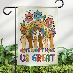 May include: A garden flag with a retro design. The flag features a rainbow, flowers, and hands of various skin tones holding the flowers. The text "Hate Won't Make Us Great" is displayed in colorful letters.