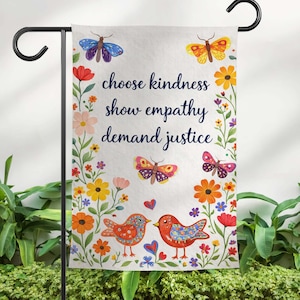 May include: A garden flag with a floral and butterfly design. The flag features the text "choose kindness, show empathy, demand justice." Two birds and several hearts are at the bottom. The flag is on a black metal stand.