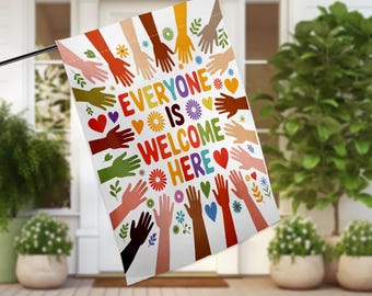 May include: A white flag with a circle of colorful hands reaching out to each other. The text "Everyone is Welcome Here" is in the center of the circle. The hands are surrounded by small hearts and flowers.