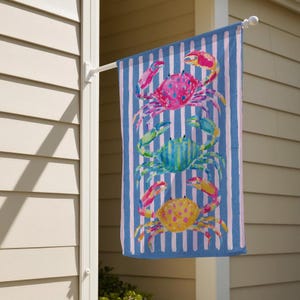 May include: A decorative outdoor flag featuring three colorful crabs stacked vertically. The crabs are painted in shades of pink, blue, and yellow, set against a blue and white striped background. The flag is hanging on a white pole.