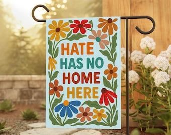 Puede incluir: Una bandera de jardín con el texto "HATE HAS NO HOME HERE" en naranja, verde y rojo. La bandera presenta un fondo azul claro con flores coloridas en tonos naranja, rojo, amarillo y azul. La bandera se muestra en un soporte de metal negro.