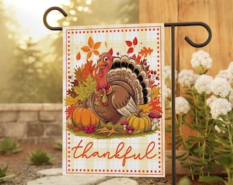 May include: Thanksgiving garden flag featuring a cartoon turkey with pumpkins and fall leaves. The flag has the word "thankful" in a decorative font. The background is a plaid pattern.