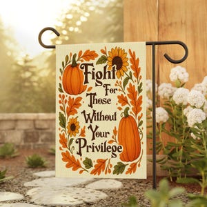 May include: A garden flag with the text "Fight For Those Without Your Privilege" surrounded by pumpkins, sunflowers, and fall leaves. The flag is beige and is hanging on a black metal stand in a garden setting.
