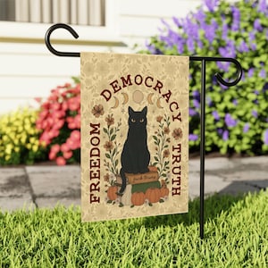 May include: A beige garden flag with a black cat illustration, surrounded by floral and celestial designs. The flag features the words "DEMOCRACY TRUTH FREEDOM" and "fuck trump". The flag is displayed on a black metal stand in a garden.