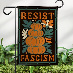 May include: A garden flag with the words "RESIST FASCISM" in orange letters. The flag features a stack of orange pumpkins, white daisies, and teal leaves against a black background. The flag is hanging on a black metal stand.