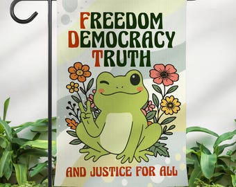 May include: A garden flag featuring a cartoon frog giving a peace sign, surrounded by flowers. The flag has the words "FREEDOM DEMOCRACY TRUTH" and "AND JUSTICE FOR ALL" in bold text. The flag is displayed on a black metal stand.