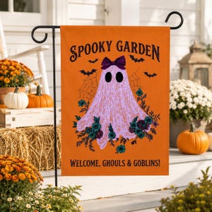 Könnte beinhalten: Eine orangefarbene Gartenflagge mit einem hellvioletten Geist mit einer dunkelvioletten Schleife, umgeben von Spinnweben und blaugrünen Blumen. Der Text lautet SPOOKY GARDEN oben und WELCOME, GHOULS & GOBLINS! unten, mit kleinen schwarzen Fledermäusen.