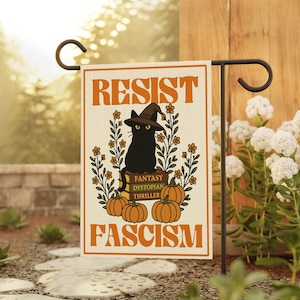 May include: A garden flag with the word "RESIST" in orange block letters. A black cat wearing a witch hat sits on books labeled "FANTASY DYSTOPIAN THRILLER." Pumpkins and flowers surround the cat. The word "FASCISM" is at the bottom.