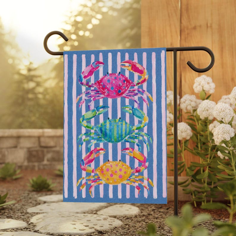 May include: A blue and white striped garden flag featuring three colorful crabs. The crabs are painted in shades of pink, green, and yellow, with detailed claws and bodies. The flag is displayed on a black metal stand in a garden setting.