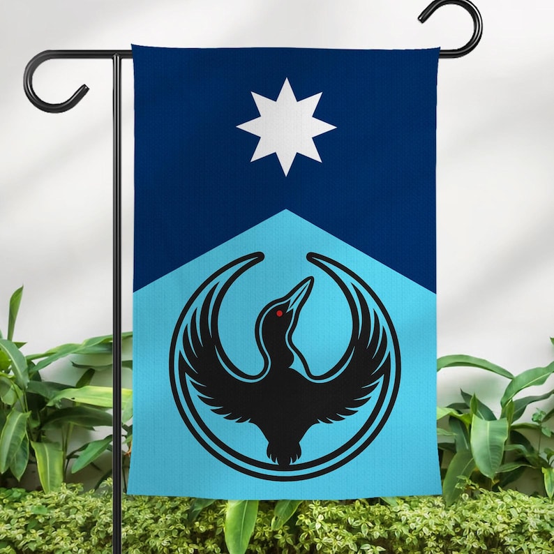 Op de afbeelding: Tuinvlag met een gestileerd zwart vogel-embleem op een blauwe en lichtblauwe achtergrond. Het ontwerp bevat een witte ster en een gestileerde vogel. De vlag hangt aan een zwarte metalen standaard.
