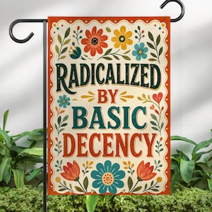 May include: A rectangular garden flag featuring a cream background with a decorative orange border. The text RADICALIZED BY BASIC DECENCY is printed in dark green and orange. The design includes colorful floral illustrations in red, yellow, and blue.