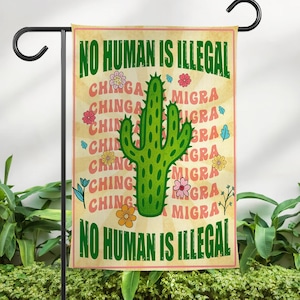 May include: A garden flag with the text "NO HUMAN IS ILLEGAL" in green, with a green cactus in the center. The flag has a beige background with the words "CHINGA" and "MIGRA" in red. Colorful flowers and leaves are scattered around the cactus.