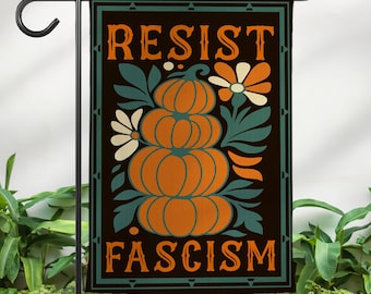 May include: A garden flag with the words "RESIST FASCISM" in orange letters. The flag features a stack of orange pumpkins, white daisies, and teal leaves against a black background. The flag is hanging on a black metal stand.