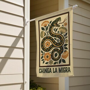 May include: A rectangular beige garden flag with a decorative snake and floral design. The snake is green and brown, with an open mouth and a long tongue. The flag has the text "CHINGA LA MIGRA" in dark green.