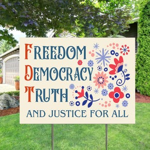 May include: A rectangular yard sign with the words "FREEDOM DEMOCRACY TRUTH AND JUSTICE FOR ALL" in red and blue lettering. The sign has floral and star designs in red, pink, and blue. The sign is on a green lawn.