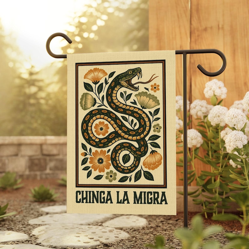 May include: A garden flag featuring a stylized illustration of a green and brown snake with an open mouth, surrounded by orange and green flowers. The text "CHINGA LA MIGRA" is printed at the bottom of the flag. The flag is displayed on a black metal stand.