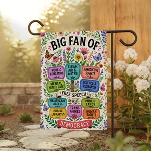 May include: A garden flag with the text "BIG FAN OF" and various phrases like "Free Speech" and "Democracy." The flag features colorful floral and butterfly designs, with phrases on signs such as "Public Education" and "Human Rights."