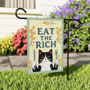 May include: A garden flag with the text "EAT THE RICH" and a cartoon cat holding a knife and fork. The flag has a light yellow background with floral accents and a green border. The flag is displayed on a black metal stand.