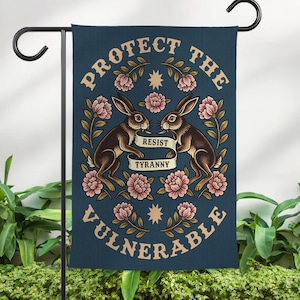 May include: A dark blue garden flag featuring two brown rabbits facing each other, with a banner reading "RESIST TYRANNY." The flag is decorated with pink flowers, gold stars, and the words "PROTECT THE VULNERABLE" in a circular design.