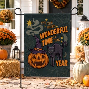 Könnte beinhalten: Eine dunkelgrüne Gartenflagge mit einem Kürbis, der einen violetten Hexenhut trägt, einer schwarzen Katze und einem Spinnennetz-Muster. Der Text lautet IT'S THE MOST WONDERFUL TIME OF THE YEAR in orangefarbenen Buchstaben mit gelben Sternen.