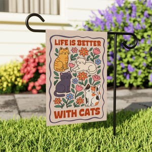 May include: A beige garden flag with the text "LIFE IS BETTER WITH CATS" in a brown border. The flag features illustrations of four cats in orange, white, black, and calico, surrounded by colorful flowers. The flag is displayed on a black metal stand.