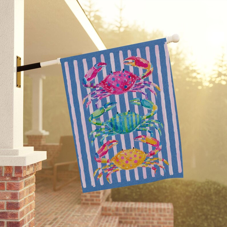May include: A decorative outdoor flag featuring three colorful crabs stacked vertically. The crabs are pink, blue, and yellow, set against a blue and white striped background. The flag is mounted on a white pole.