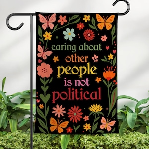 May include: A black garden flag with colorful floral and butterfly designs. The flag features the text "caring about other people is not political" in various colors. The flag is displayed on a black metal stand.