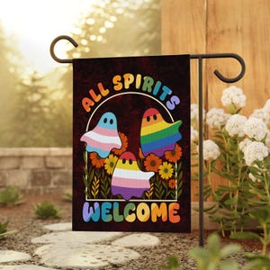 May include: A garden flag with the text "ALL SPIRITS WELCOME" and three cartoon ghosts in front of flowers. The ghosts are colored with the transgender flag, the rainbow pride flag, and the non-binary flag. The flag is on a black metal stand.