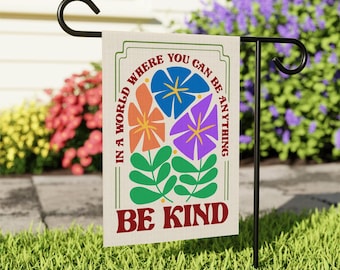 May include: A garden flag with the text "In a world where you can be anything, be kind." The flag features colorful flower illustrations in orange, blue, and purple, with green leaves. The flag is displayed on a black metal stand in a garden setting.