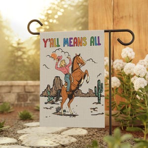 May include: A garden flag with a retro illustration of a cowgirl riding a rearing horse in a desert landscape. The flag reads "Y'ALL MEANS ALL" in colorful letters. The flag is displayed on a black metal stand.