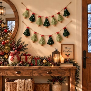 K&ouml;nnte beinhalten: Zwei horizontale Girlanden mit abwechselnd hell- und dunkelgr&uuml;nen geh&auml;kelten Weihnachtsbaum-Anh&auml;ngern. Jeder Baum ist mit einer kleinen roten Schleife versehen und mit wei&szlig;en Punkten verziert, aufgeh&auml;ngt an einer d&uuml;nnen braunen Schnur.