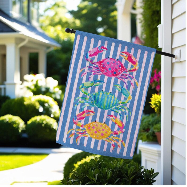 May include: A blue and white striped garden flag features three colorful crabs. The crabs are pink, blue, and yellow with polka dots and are set against a blue and white striped background. The flag is hanging outside a house.