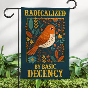May include: A navy blue garden flag with a bird illustration and the text "RADICALIZED BY BASIC DECENCY" in gold lettering. The bird is orange and white, surrounded by colorful floral and leaf designs.