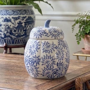 May include: A white and blue ceramic pumpkin-shaped jar with a lid. The jar features a blue floral pattern and a blue stem. The jar is on a wooden table, with a large blue and white planter in the background.