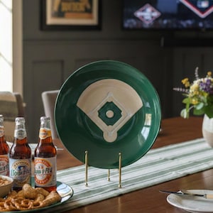 May include: A green ceramic plate with a baseball field design, displayed on a gold stand. The table setting includes beer bottles, snacks, and a green and white striped runner, creating a sports-themed atmosphere.