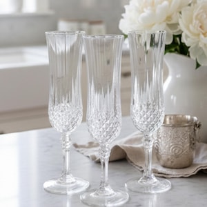 May include: Three clear crystal champagne flutes with diamond-cut bases and vertical ribbed detailing. The flutes are displayed on a marble surface. A white jug of flowers and a silver container are in the background.