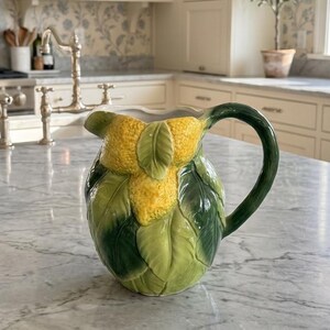 May include: A ceramic pitcher with a lemon and leaf design. The pitcher features a yellow lemon cluster and green leaves, with a green handle. The pitcher is sitting on a marble countertop in a kitchen setting.