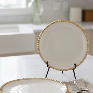Pode incluir: Pratos de cerâmica branca com borda dourada com contas. Um prato é exibido em um suporte de metal preto, enquanto outros são empilhados em uma mesa. Um garfo e uma colher de prata repousam sobre uma toalha de linho bege.