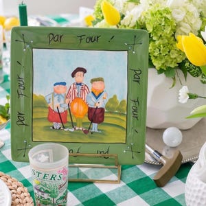 May include: A green square decorative plate with a golf-themed illustration. The plate features a painting of three golfers on a green course, with the words "Par Four!" around the border. A golf ball and putter are also visible.