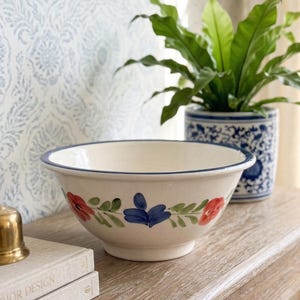 May include: A cream-colored ceramic bowl with a blue rim and floral design. The bowl features red and blue flowers with green leaves. A gold bell and a potted plant are in the background.
